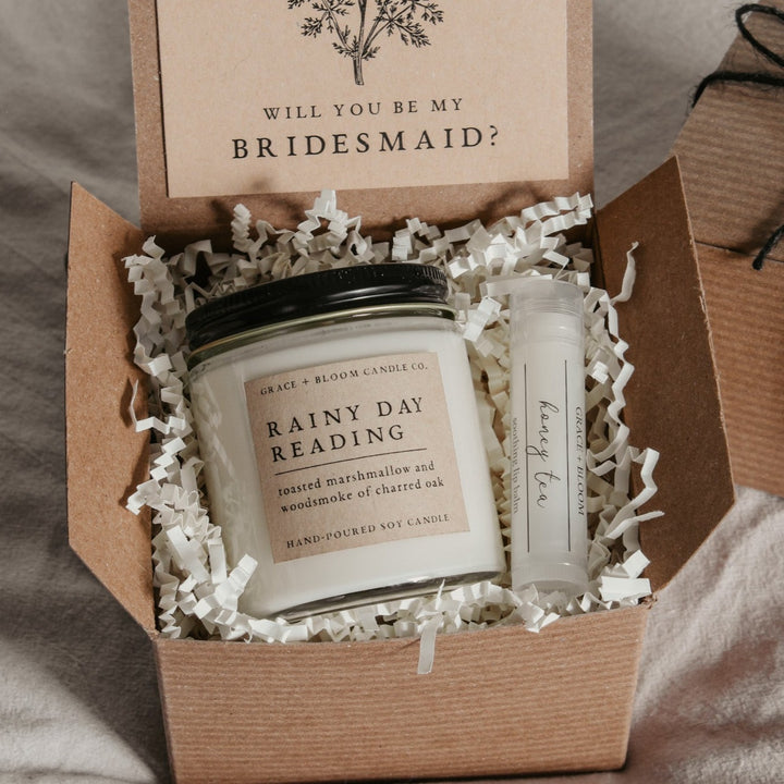 A gift box opened to reveal a scented candle and a small container of lip balm, labeled 'Rainy Day Reading' and 'Honey Tea', respectively. The box is decorated with a card with a vintage floral illustration and the text 'Will you be my bridesmaid?' is visible.