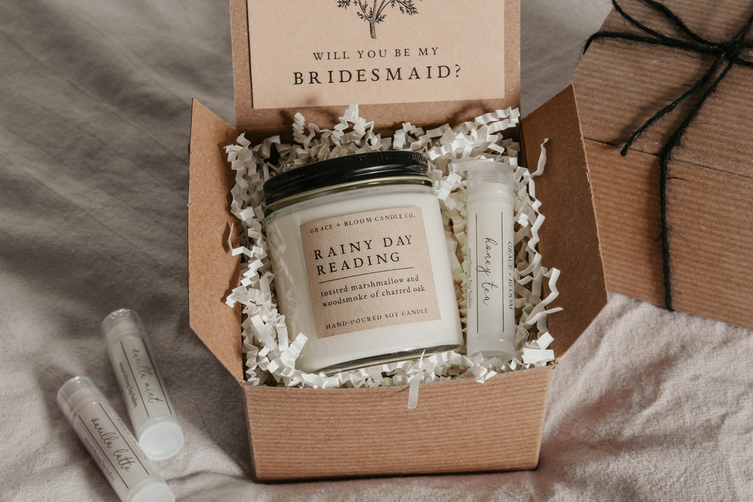 A gift box opened to reveal a scented candle and a small container of lip balm, labeled 'Rainy Day Reading' and 'Honey Tea', respectively. The box is decorated with a card with a vintage floral illustration and the text 'Will you be my bridesmaid?' is visible.