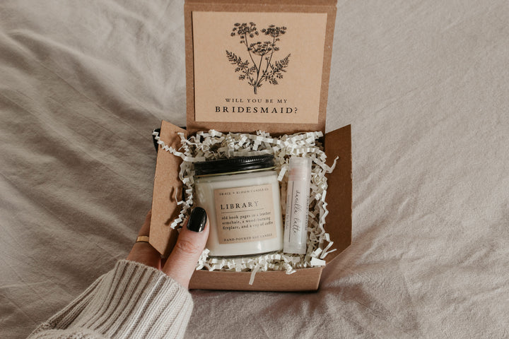 A gift box opened to reveal a scented candle and a small container of lip balm, labeled 'Library' and 'Vanilla Latte', respectively. The box is decorated with a card with a vintage floral illustration and the text 'Will you be my bridesmaid?' is visible.