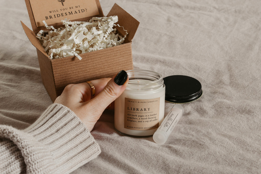 a hand holding a small candle and a lip balm with the black metal candle lid and an open kraft gift box in the background