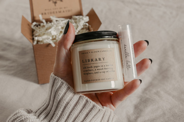 hands holding a small candle with a kraft label and a black metal lid and a lip balm with an open kraft gift box in the background