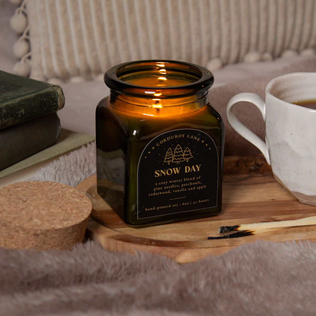 Green glass candle labeled 'Snow Day' on a wooden surface with a cup and books in the background.