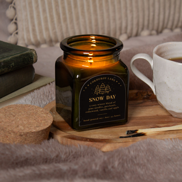 Green glass candle labeled 'Snow Day' on a wooden surface with a cup and books in the background.