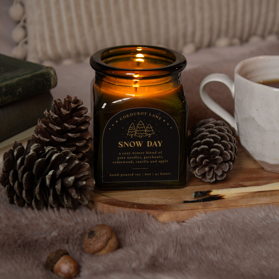 Candle labeled 'Snow Day' with pinecones and a mug on a wooden board.