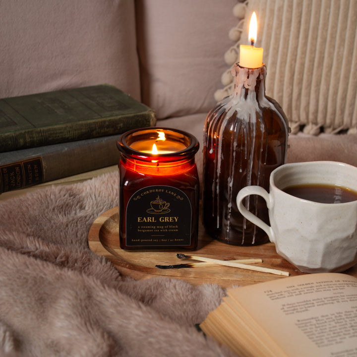 Candle labeled 'Earl Grey' on a wooden tray with a cup of coffee, book, and decorative bottle.