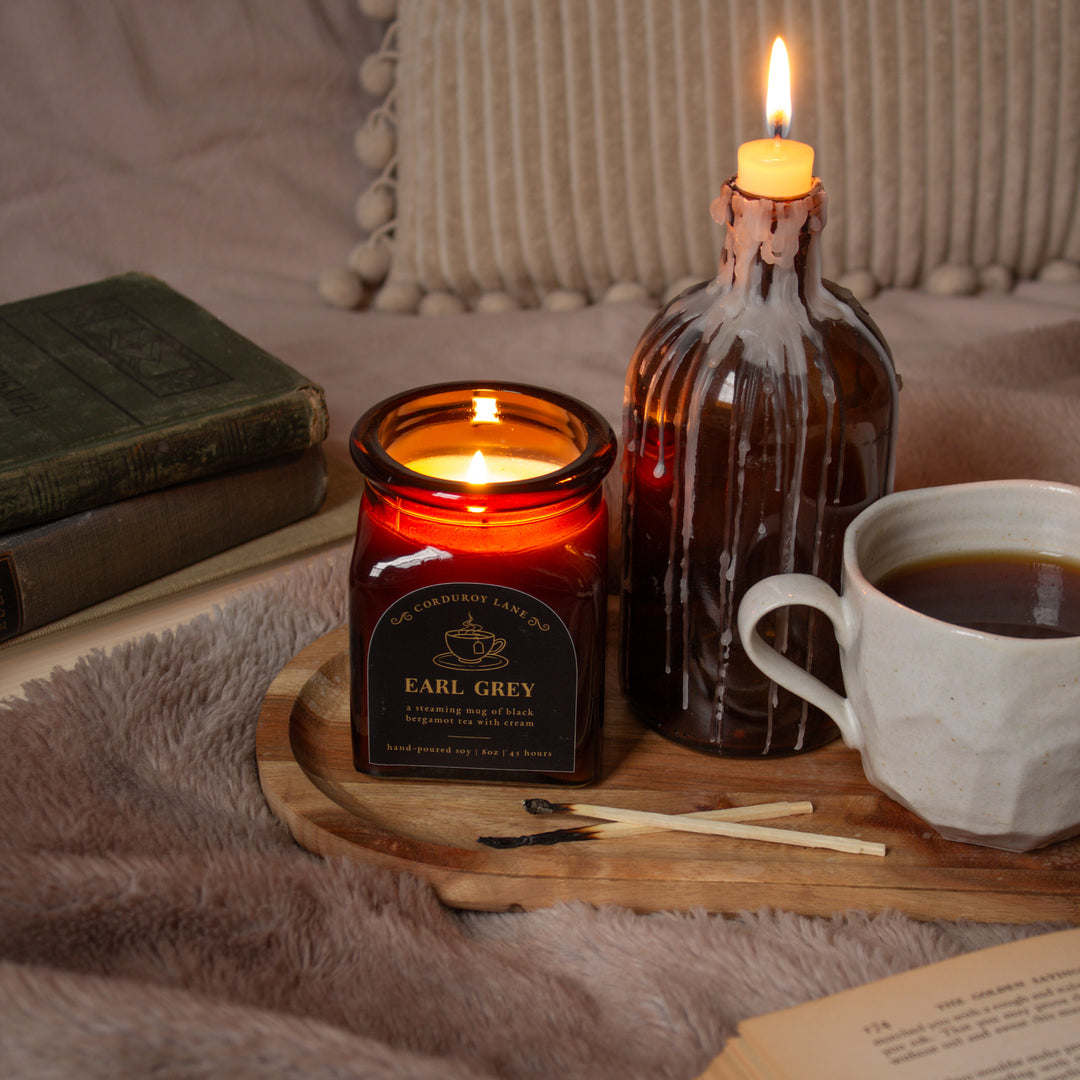 Two lit candles, one in a square amber glass jar labeled 'Earl Grey' and the other in a decorative bottle, on a wooden tray with a cup of coffee and books in the background.