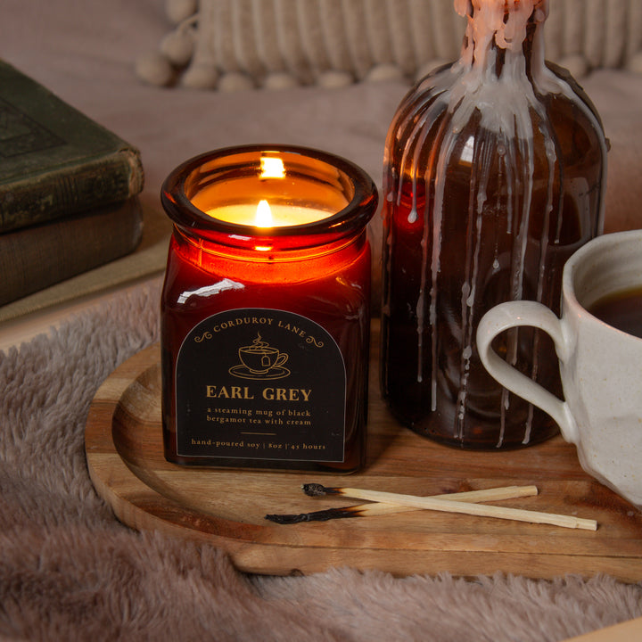 Candle labeled 'Earl Grey' on a wooden tray with a bottle and cup in the background.