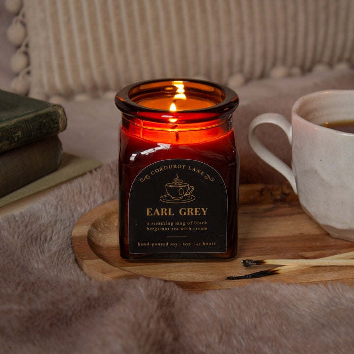 Candle labeled 'Earl Grey' on a wooden coaster with a cup of tea and books in the background.