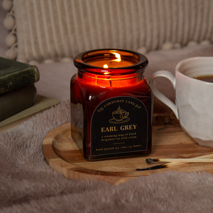 Candle labeled 'Earl Grey' on a wooden tray with a cup of tea and books in the background.