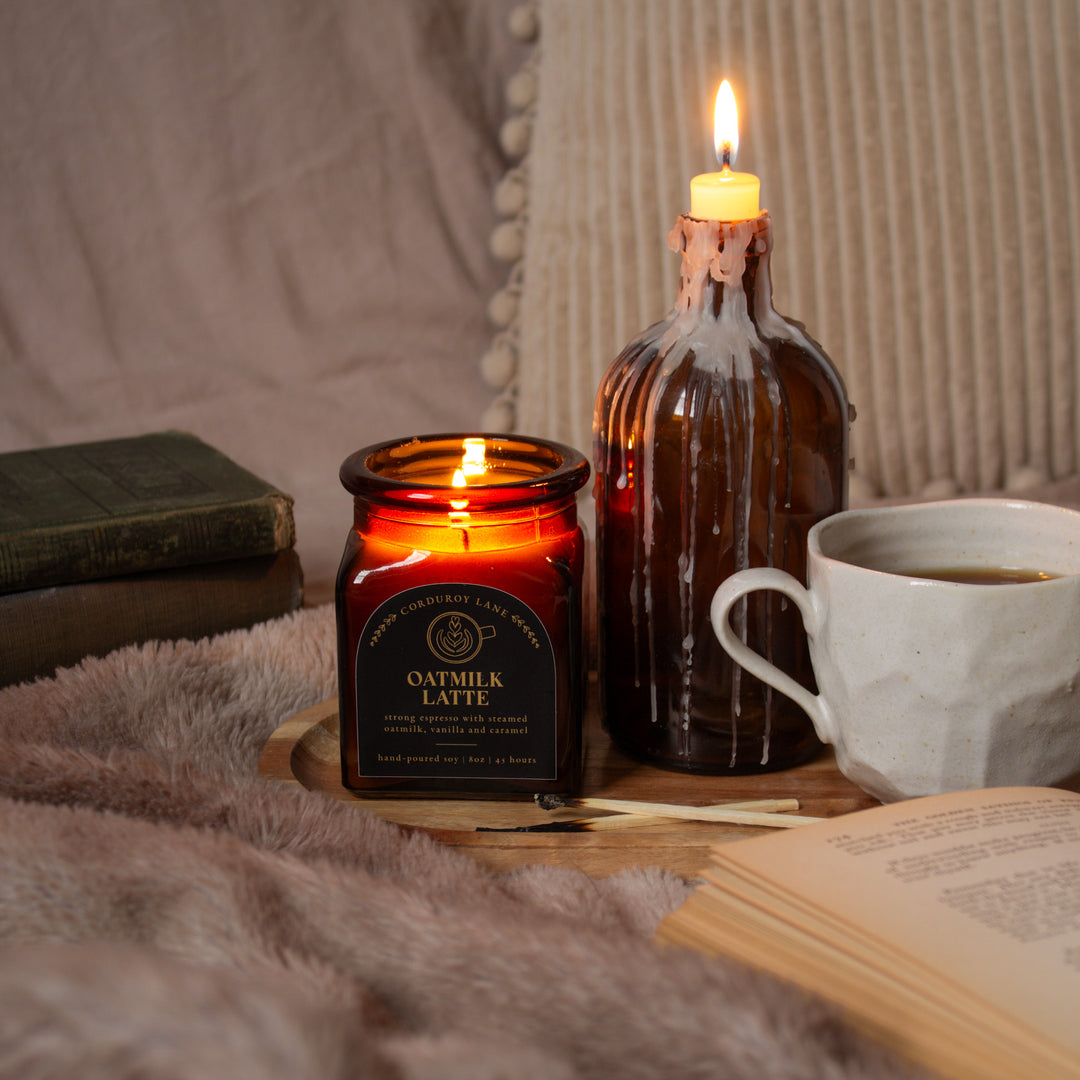 Two lit candles in glass jars, a cup of coffee, and an open book on a textured surface.