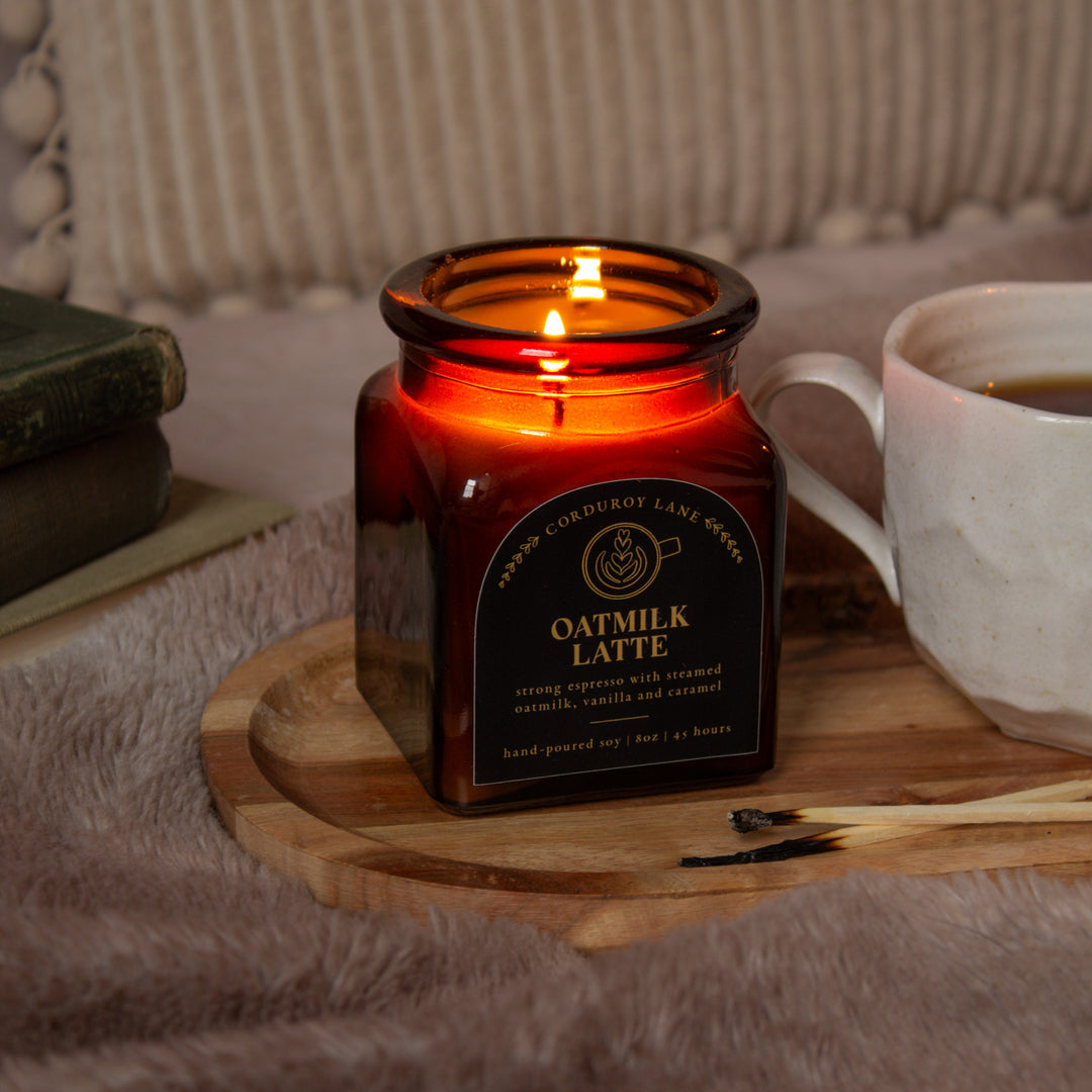 Candle in a square amber apothecary jar labeled 'Oatmilk Latte' on a wooden tray with a cup of coffee and books in the background.