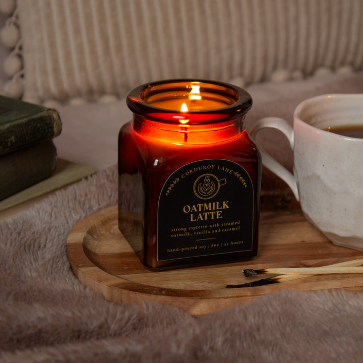 Candle in a square amber apothecary jar labeled 'Oatmilk Latte' on a wooden tray with a cup of coffee and books in the background.