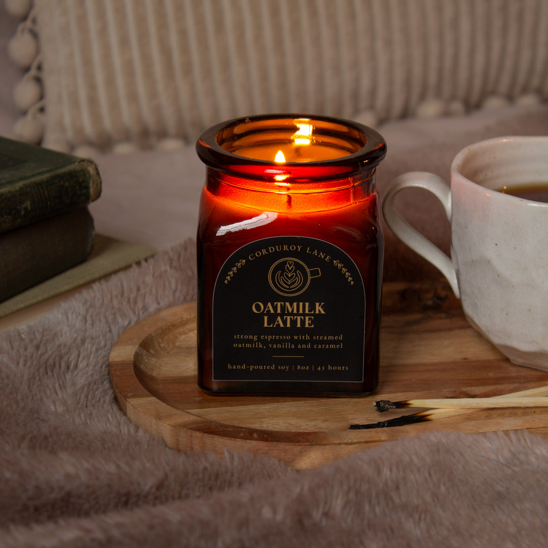 Candle labeled 'Oatmilk Latte' on a wooden coaster with a cup of coffee and books in the background.