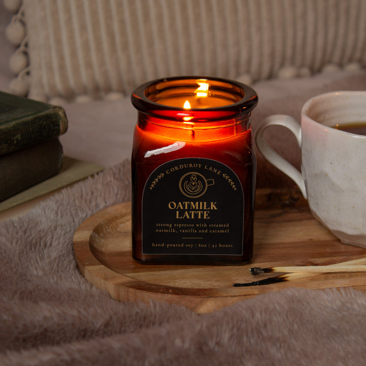 Candle labeled 'Oatmilk Latte' on a wooden coaster with a cup of coffee and books in the background.