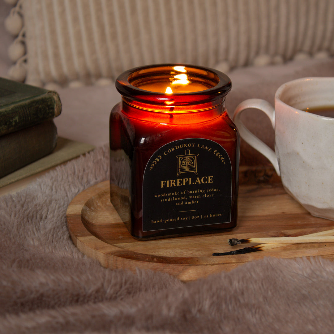 Lit candle in a square amber apothecary  jar labeled 'Fireplace' on a wooden coaster with a cup of coffee and books in the background.