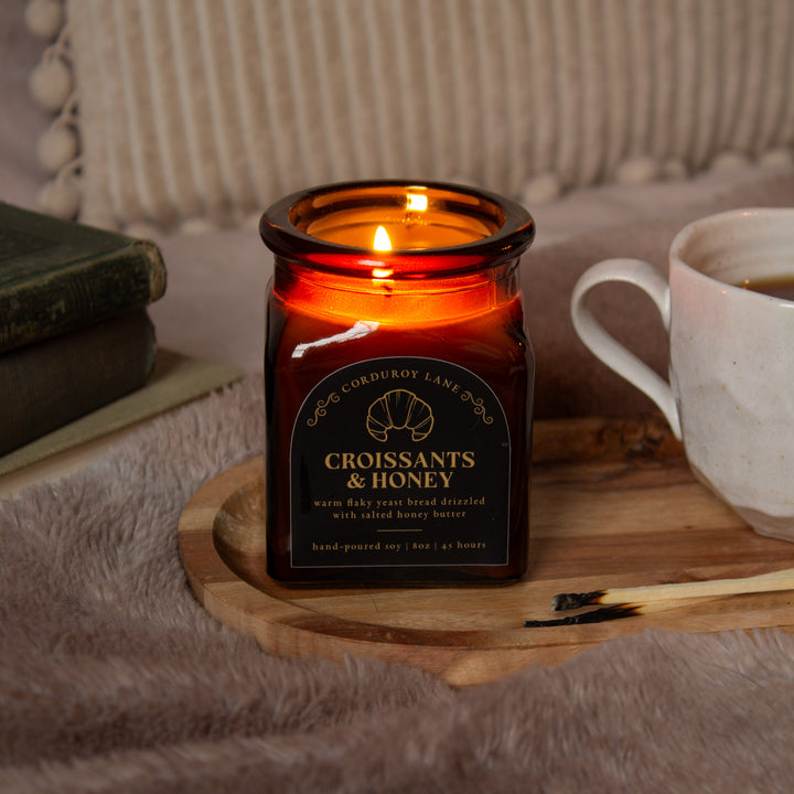 Candle in a decorative jar with a label, next to a mug on a wooden surface.