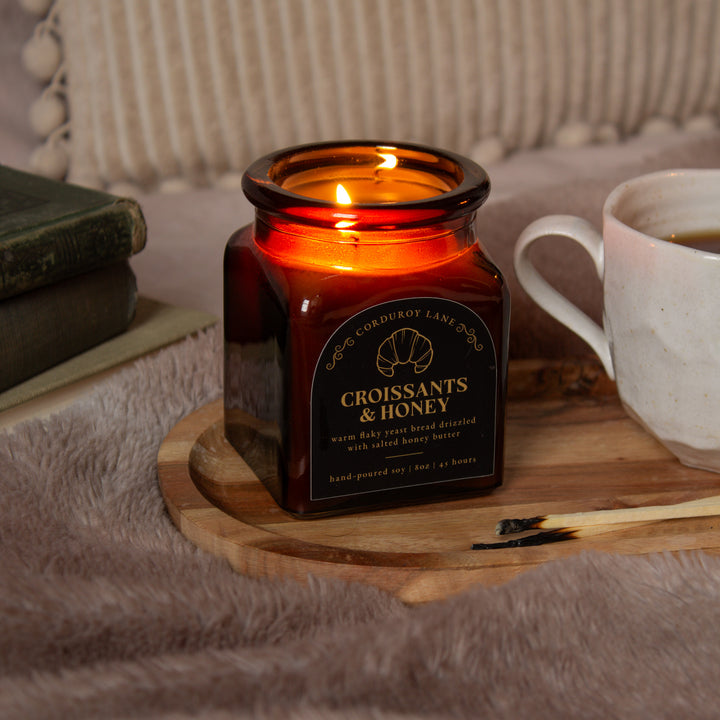 Candle in an square amber apothecary jar labeled 'Croissants & Honey' on a wooden tray with a cup and books in the background.