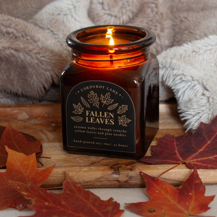 Candle labeled 'Fallen Leaves' with autumn leaves around on a wooden surface.