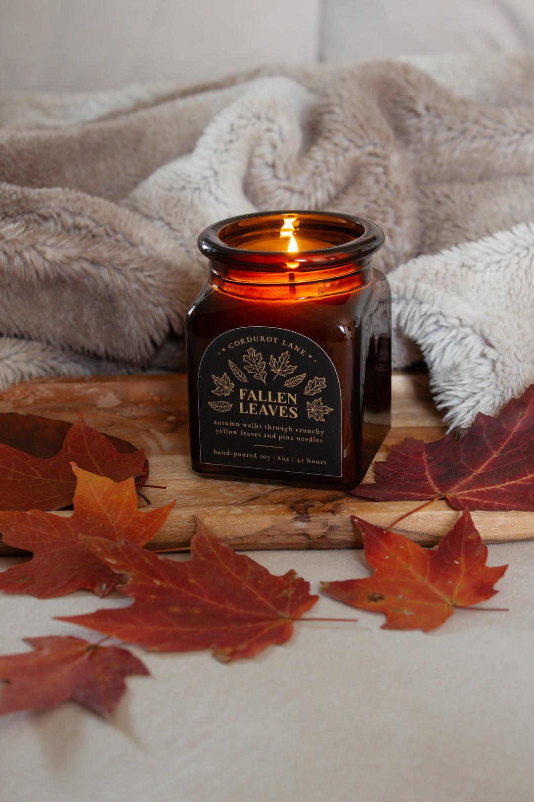 Candle labeled 'Fallen Leaves' on a wooden surface with autumn leaves and a blanket in the background