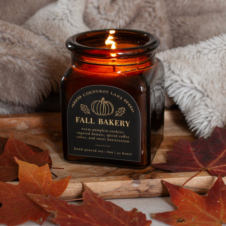 Candle in an amber jar labeled 'Fall Bakery' with autumn leaves and a cozy background