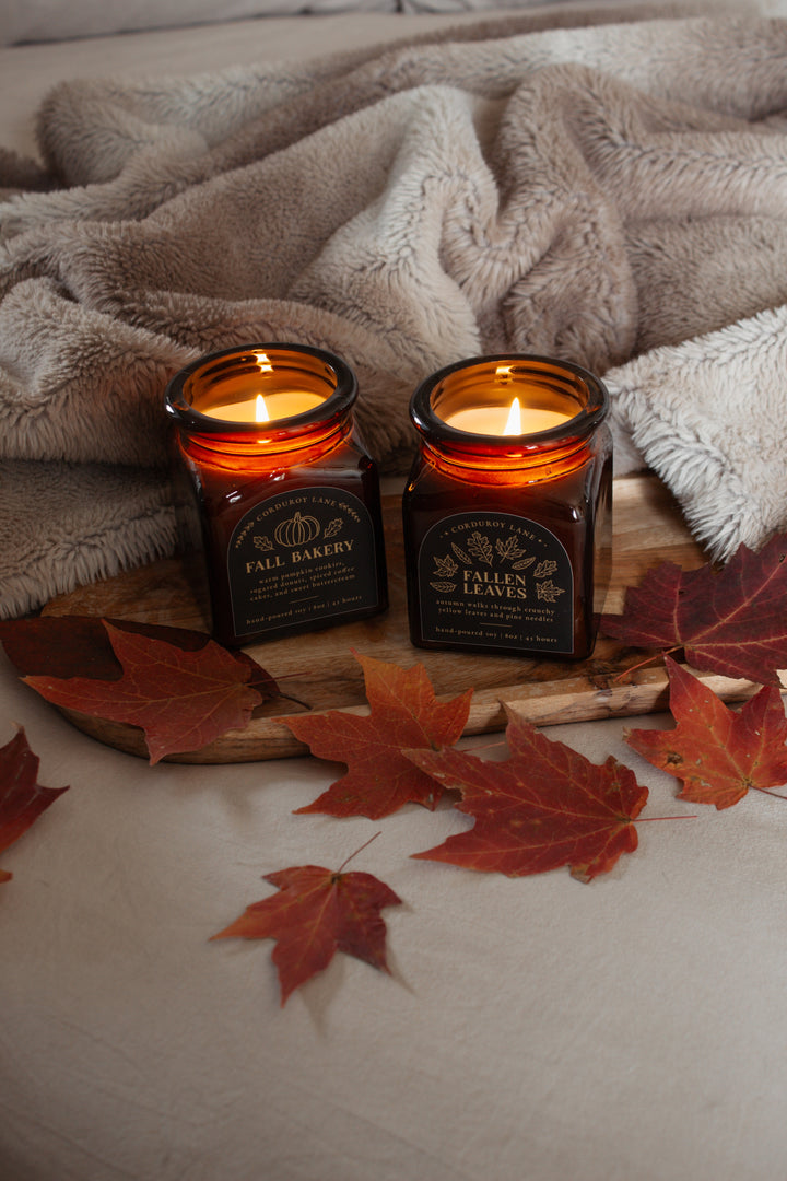 Two lit candles in amber glass jars on a wooden board with autumn leaves and a blanket.