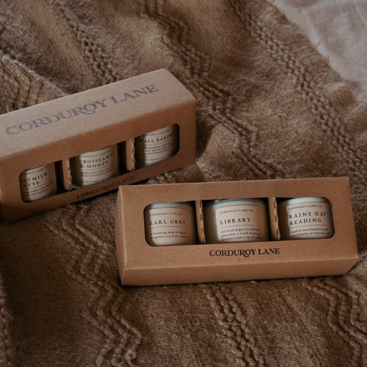 Two cardboard boxes containing small container candles labeled 'Corduroy Lane' on a textured brown surface.