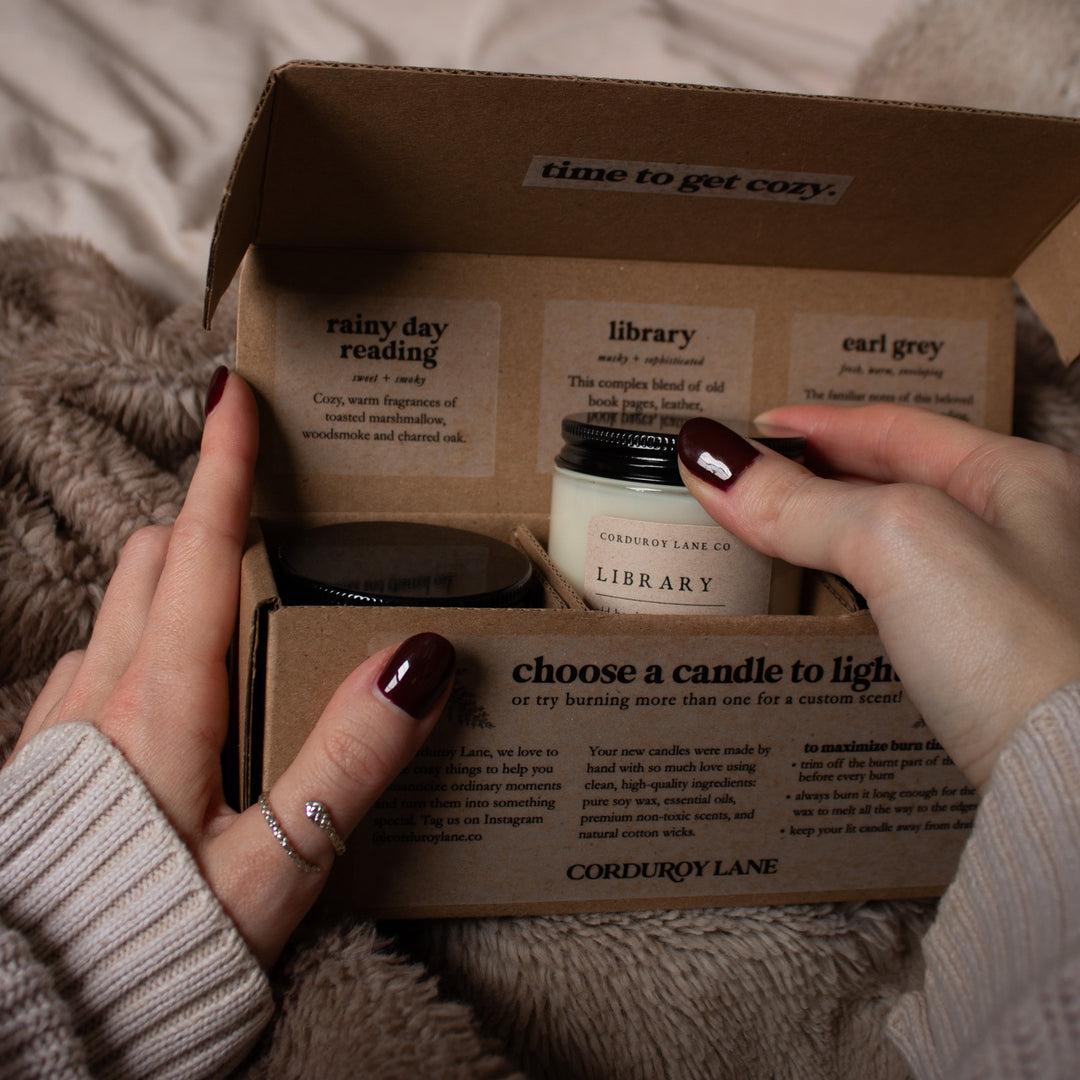 Open cardboard box with candles labeled 'Library' and 'Earl Grey', held by a person.