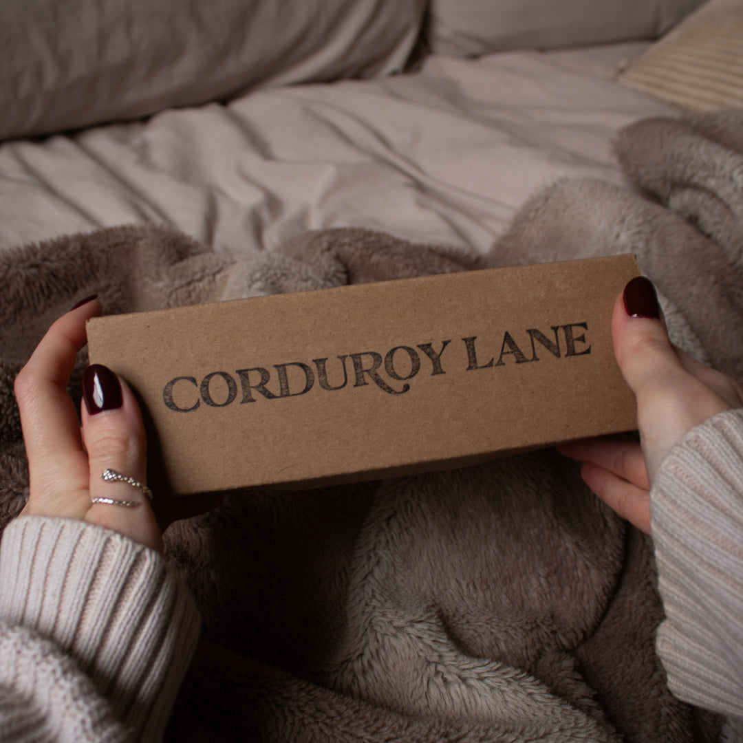 Person holding a cardboard box with 'Corduroy Lane' branding on a soft, textured surface.