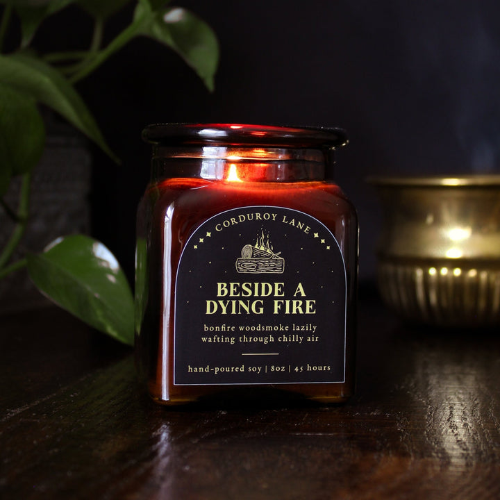 Beside a Dying Fire: a cozy fall candle from Corduroy Lane in an amber glass square apothecary jar with a cork lid and a black and gold label, scented in a warm, smoky blend of maple, clove, and earthy-green undertones of moss, patchouli, and vetiver