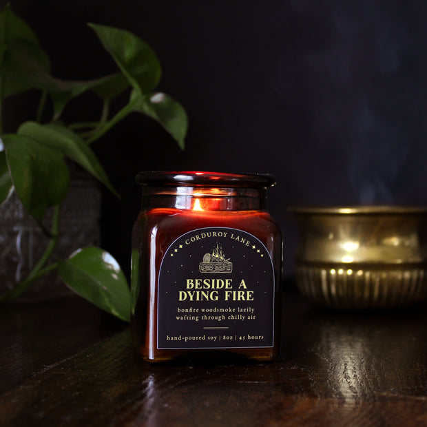 Beside a Dying Fire: a cozy fall candle from Corduroy Lane in an amber glass square apothecary jar with a cork lid and a black and gold label, scented in a warm, smoky blend of maple, clove, and earthy-green undertones of moss, patchouli, and vetiver