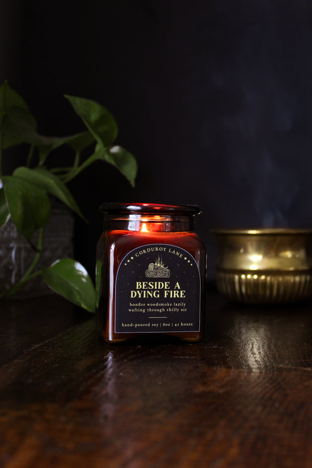 Beside a Dying Fire: a cozy fall candle from Corduroy Lane in an amber glass square apothecary jar with a cork lid and a black and gold label, scented in a warm, smoky blend of maple, clove, and earthy-green undertones of moss, patchouli, and vetiver