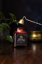 Beside a Dying Fire: a cozy fall candle from Corduroy Lane in an amber glass square apothecary jar with a cork lid and a black and gold label, scented in a warm, smoky blend of maple, clove, and earthy-green undertones of moss, patchouli, and vetiver