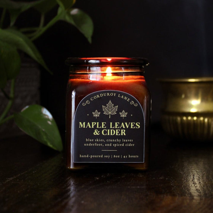 Maple Leaves and Cider: a cozy candle from Corduroy Lane in an amber glass square apothecary jar with a cork lid and a black and gold label, scented in a fresh, fruity blend of crunchy dried leaves and spiced apple cider for lovers of all things autumnal