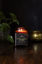 Maple Leaves and Cider: a cozy fall candle from Corduroy Lane in an amber glass square apothecary jar with a cork lid and a black and gold label, scented in a fresh, fruity blend of crunchy dried leaves and spiced apple cider for lovers of all things autumnal