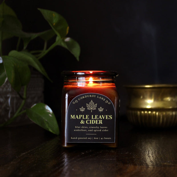 Maple Leaves and Cider: a cozy fall candle from Corduroy Lane in an amber glass square apothecary jar with a cork lid and a black and gold label, scented in a fresh, fruity blend of crunchy dried leaves and spiced apple cider for lovers of all things autumnal