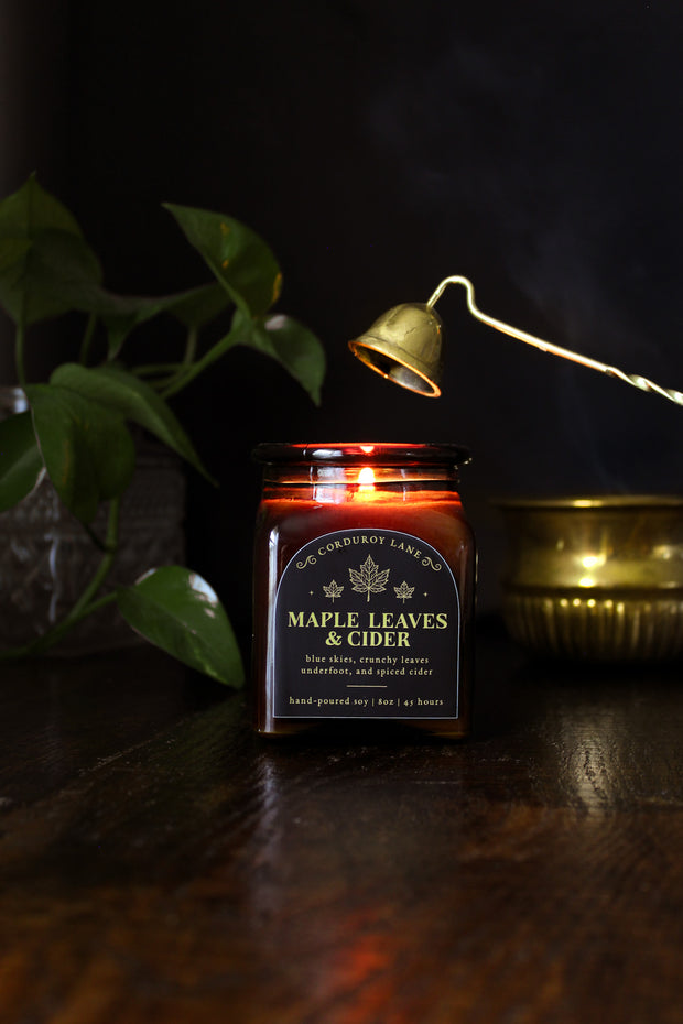 Maple Leaves and Cider: a cozy fall candle from Corduroy Lane in an amber glass square apothecary jar with a cork lid and a black and gold label, scented in a fresh, fruity blend of crunchy dried leaves and spiced apple cider for lovers of all things autumnal