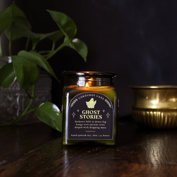 Ghost Stories: a spooky halloween candle from Corduroy Lane in a green glass square apothecary jar with a cork lid and a black and gold label, scented in a refreshing, masculine blend of misty ozone and woodlands with lavender, mint, and juniper for lovers of all things cozy and spooky