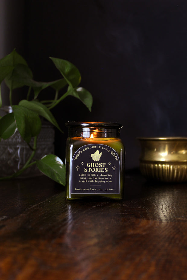 Ghost Stories: a spooky halloween candle from Corduroy Lane in a green glass square apothecary jar with a cork lid and a black and gold label, scented in a refreshing, masculine blend of misty ozone and woodlands with lavender, mint, and juniper for lovers of all things cozy and spooky