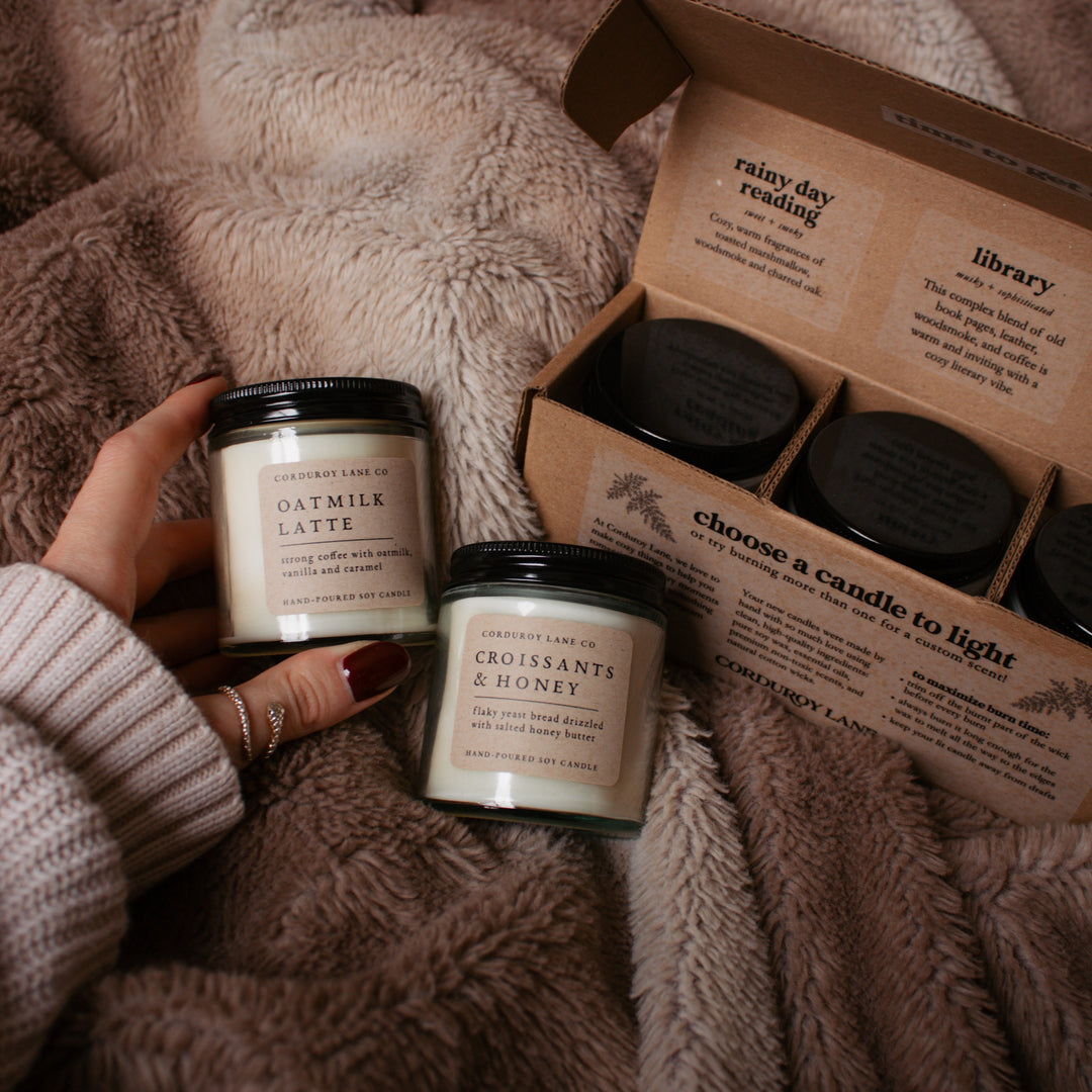 Two candles held by a hand on a textured surface with an open box of more candles in the background.