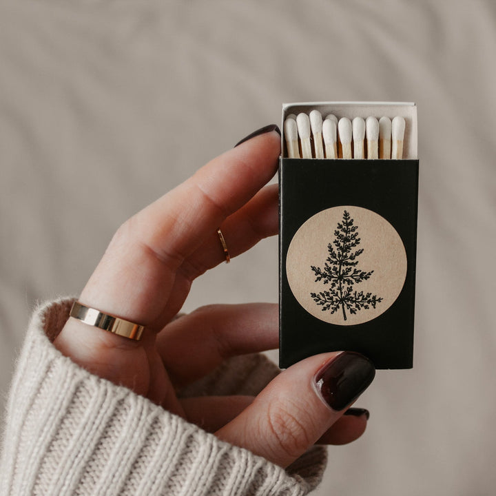 a hand holding a partially open black matchbox with white-tipped matches inside. A round sticker with a hand-drawn fern is on the front of the matchbox.