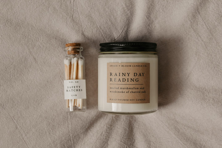 a candle labeled "Rainy Day Reading" next to a glass bottle of safety matches