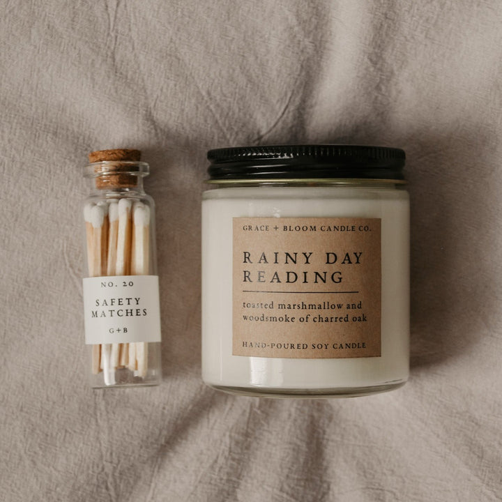 a candle labeled "Rainy Day Reading" next to a glass bottle of safety matches