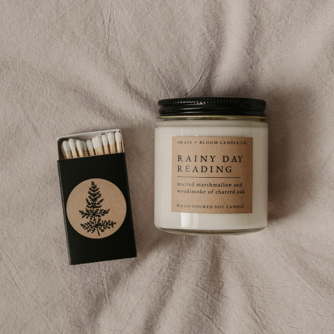 a candle labeled Rainy Day Reading with a matchbox beside it