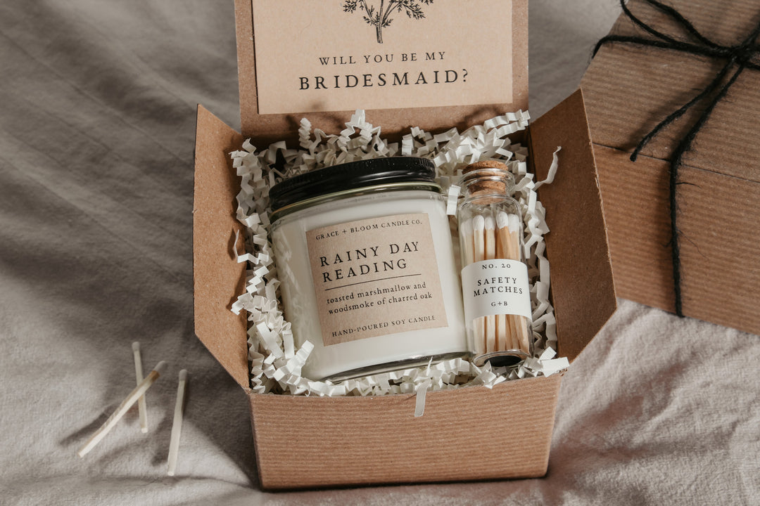 An open gift box displaying a bridesmaid proposal gift set with a scented soy candle, a custom card, and a glass bottle of matches, all themed dark academia.