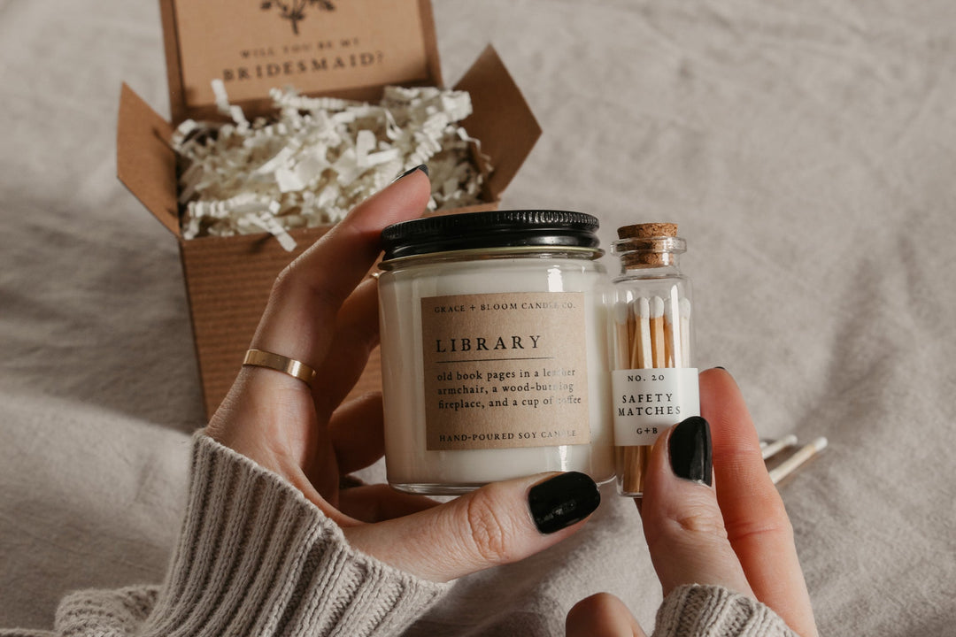 hands holding a small candle with a kraft label and a black metal lid and a glass bottle of matches with an open kraft gift box in the background