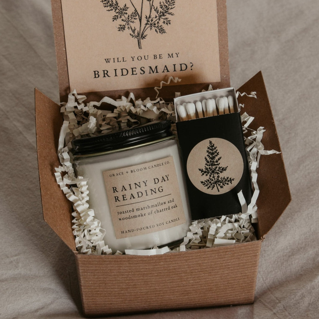 An open gift box displaying a bridesmaid proposal gift set with a scented soy candle, a custom card, and a matchbox, all themed dark academia.