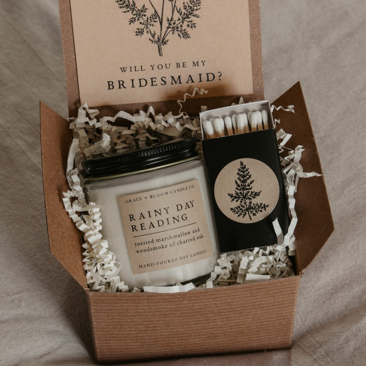 An open gift box displaying a bridesmaid proposal gift set with a scented soy candle, a custom card, and a matchbox, all themed dark academia.