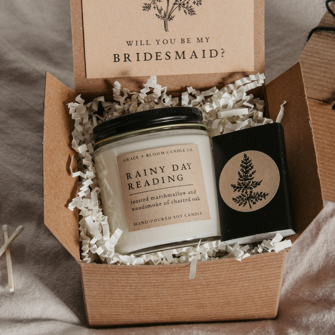 An open gift box displaying a bridesmaid proposal gift set with a scented soy candle, a custom card, and a matchbox, all themed dark academia.