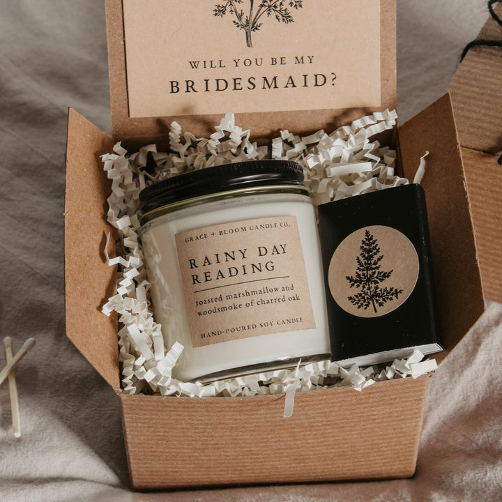 An open gift box displaying a bridesmaid proposal gift set with a scented soy candle, a custom card, and a matchbox, all themed dark academia.