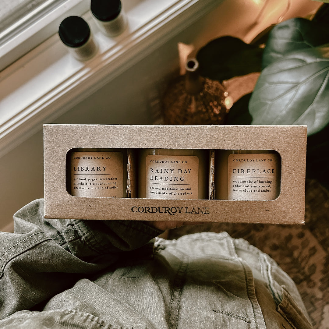 Three candles in a cardboard box labeled 'Corduroy Lane' on a surface with a blurred background.